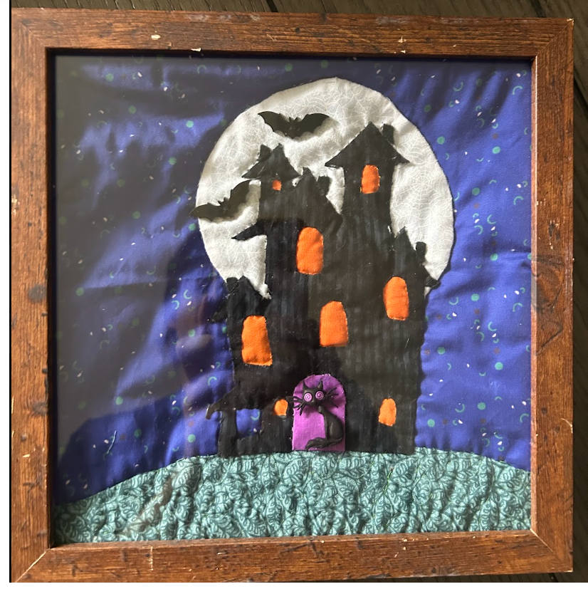 A framed handmade quilted art piece depicting a spooky haunted house at night. The quilt features a black house with orange glowing windows, a purple door, a green hill, a white full moon, and bats against a blue starry sky, all enclosed in a rustic brown wooden frame.