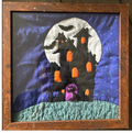 A framed handmade quilted art piece depicting a spooky haunted house at night. The quilt features a black house with orange glowing windows, a purple door, a green hill, a white full moon, and bats against a blue starry sky, all enclosed in a rustic brown wooden frame.
