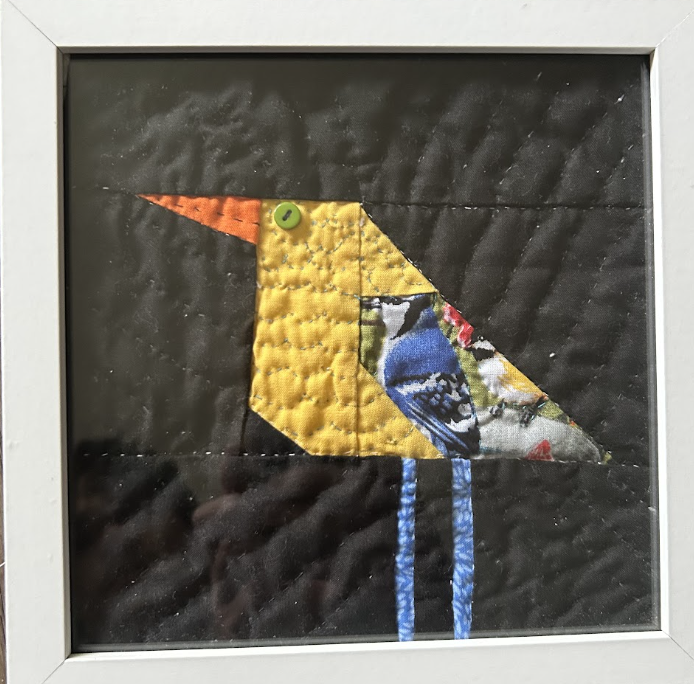 Geometric patchwork bird quilted appliqué wall art with green button eye on dark charcoal background in white frame