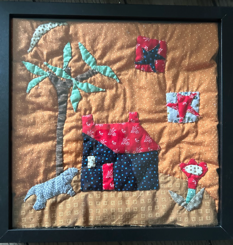 Framed folk art quilted wall art with house, palm tree, dog, cat and red flowers on golden background in black frame