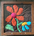 Framed quilted flower wall art with bold orange daisy, magenta center, and teal leaves on dark background in wood frame