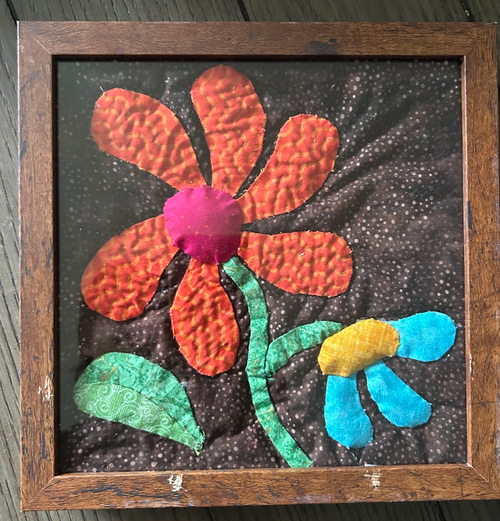 Framed quilted flower wall art with bold orange daisy, magenta center, and teal leaves on dark background in wood frame