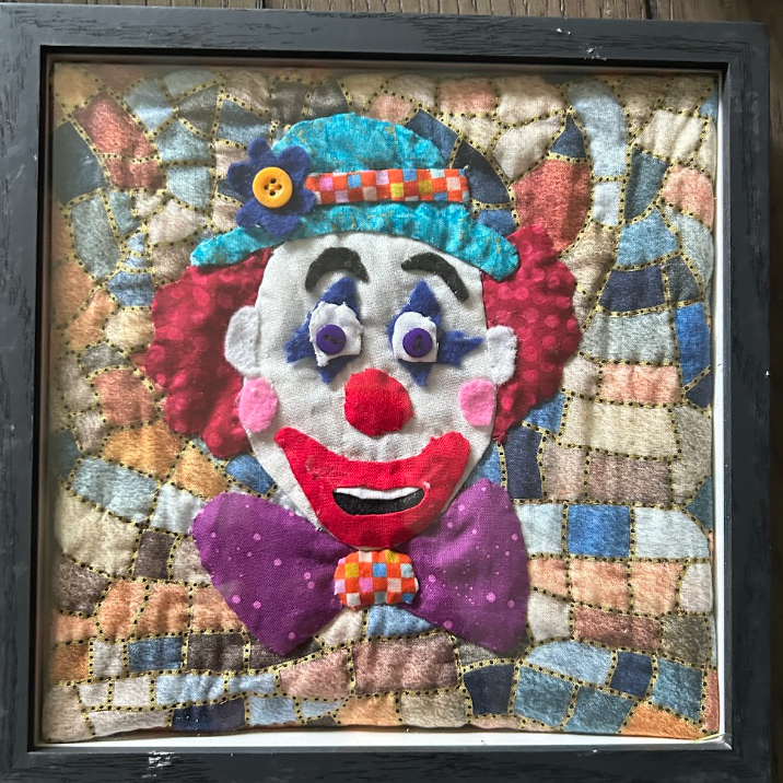 Framed quilted clown portrait wall art with teal hat, red hair, purple bow tie and patchwork background in black frame