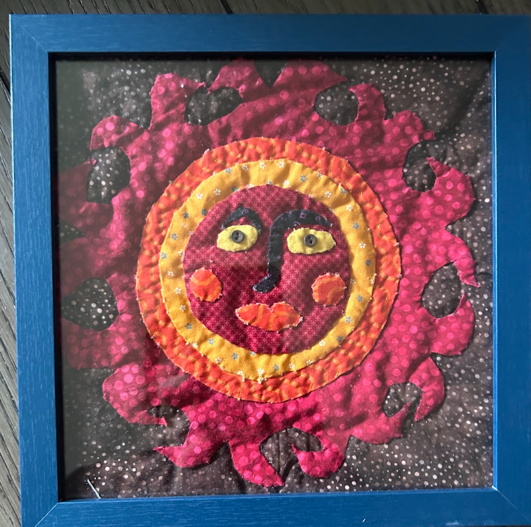 Framed quilted sun face wall art with crimson, orange, gold and magenta fabric rays on dark background in cobalt blue frame