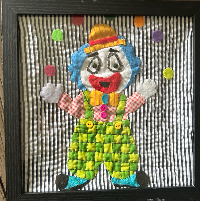 Framed quilted juggling clown wall art with green checkered overalls, blue hair, striped hat and colorful polka dots on black and white striped background