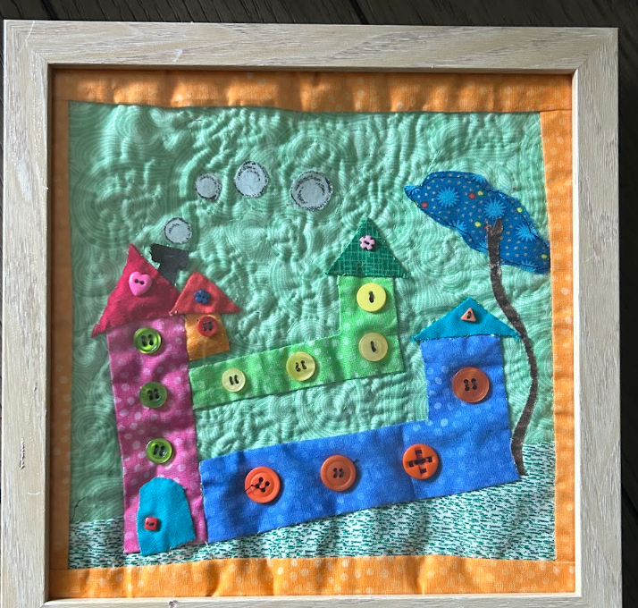 Framed quilted button house village wall art with pink, green and teal houses, blue tree and colorful button details on aqua background