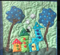 Handmade quilted whimsical village wall art with colorful houses, blue polka-dot trees, button details and a cat on swirling green background