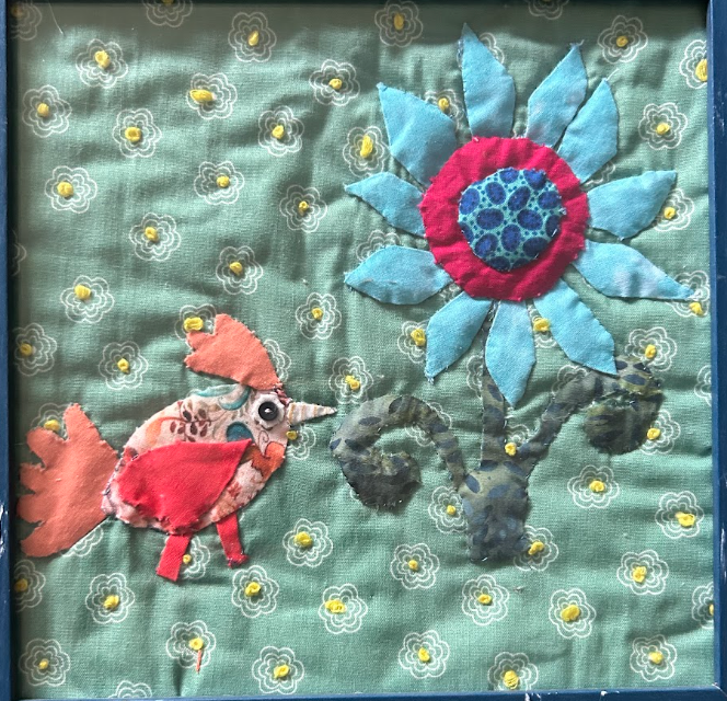 Framed quilted wall art with orange bird looking up at bold blue daisy flower on sage green floral background in dark blue frame
