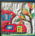 Framed quilted red house wall art with smiling faces, yellow door, blue windows, autumn tree with colorful leaves and a bird on natural linen background