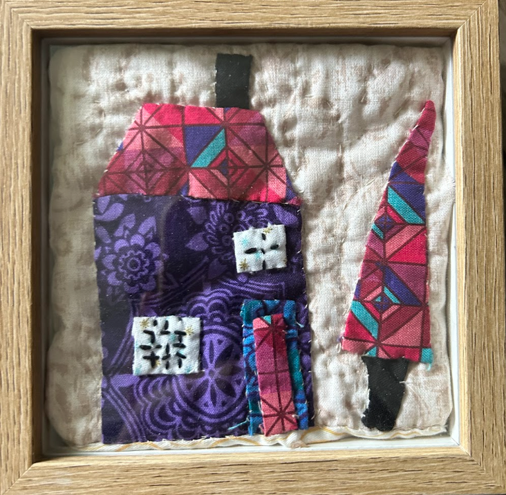 Framed quilted wall art with tall purple house, red geometric patchwork roof and matching Christmas tree on natural linen background in wood frame