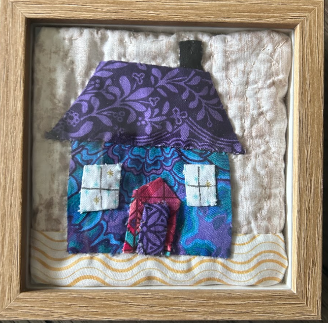 Framed quilted cottage wall art with purple floral roof, teal blue walls, red arched door and star-accented windows on natural linen background in wood frame