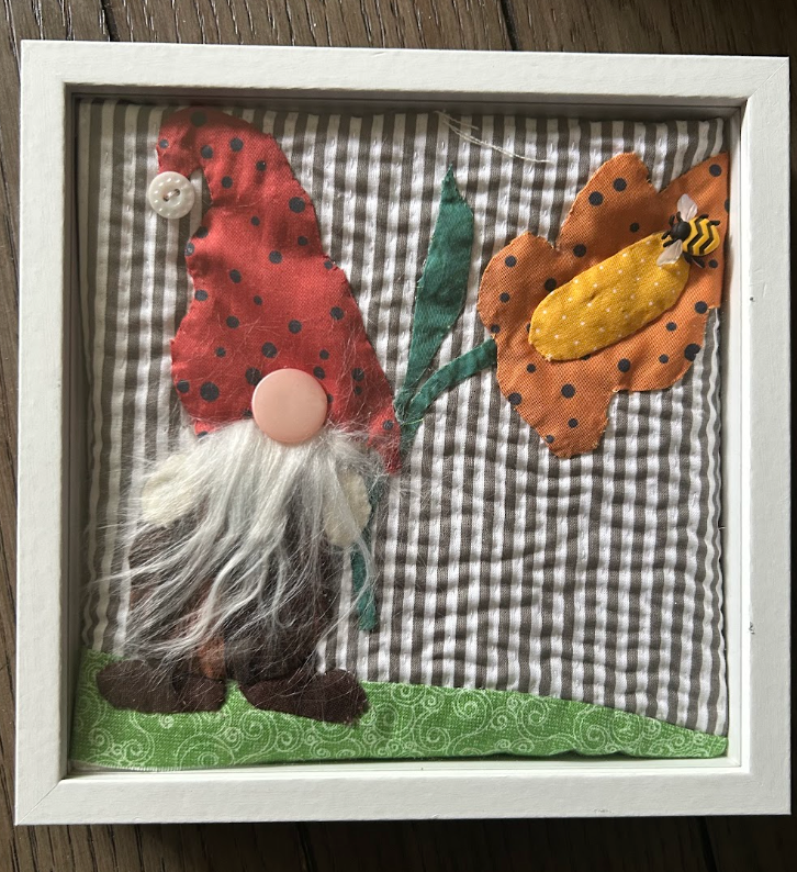 Framed quilted garden gnome wall art with red polka-dot hat, white yarn beard, orange flower and bee on grey gingham background in white frame